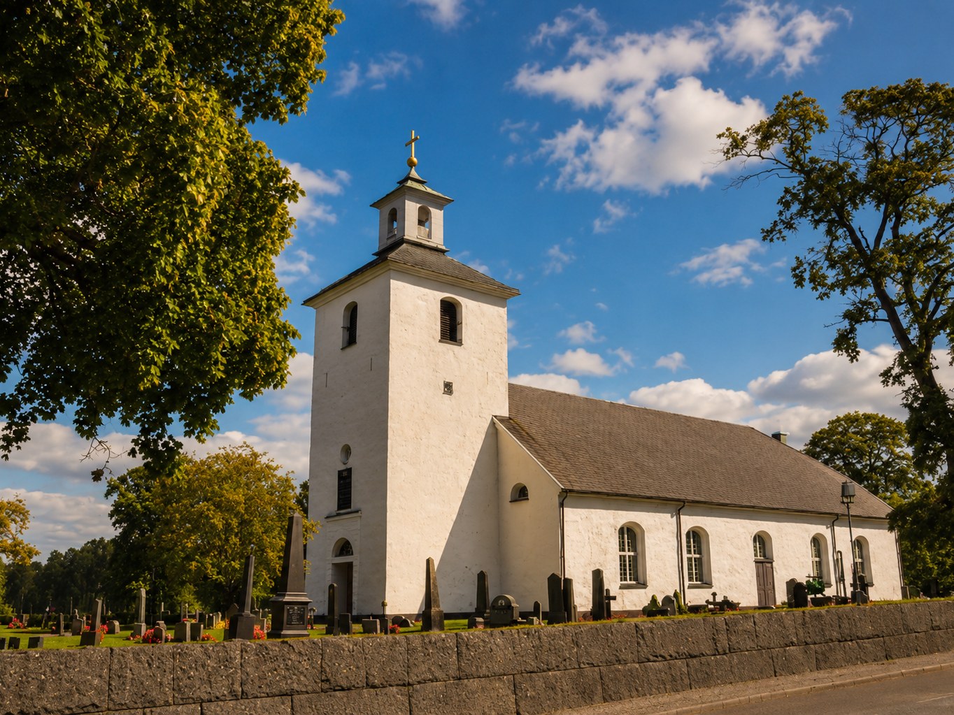 Skatelövs kyrka — vit landskyrka med klocktorn omgiven av kyrkogård och lövträd. Foto: Barbro Thörn.
